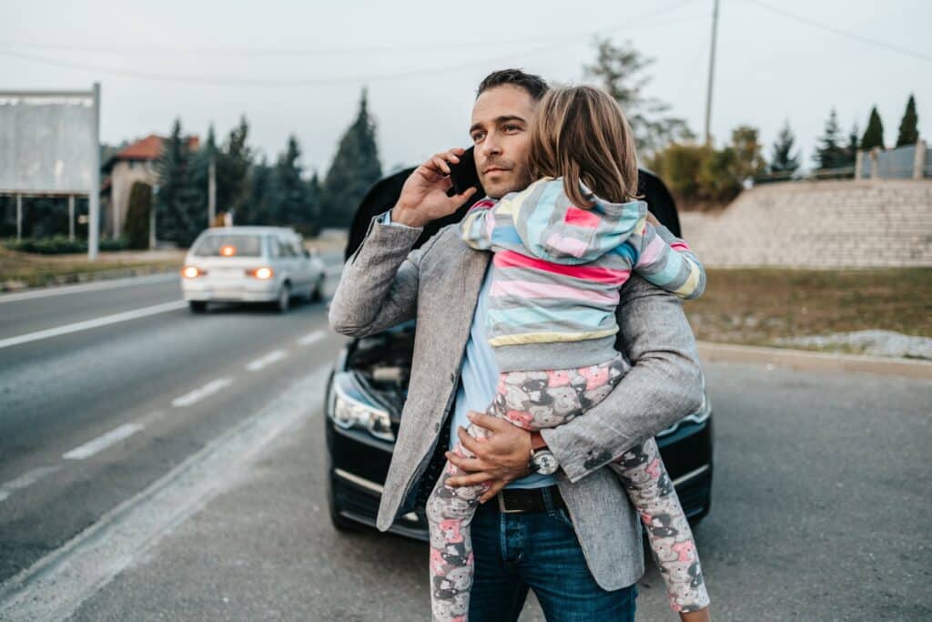Man holding a child and on the phone with two cars in the background