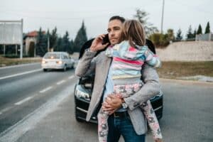 Man holding a child and on the phone with two cars in the background