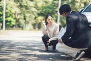 Man holding clip board taking report on car accident and woman involved in accident sitting in front of car
